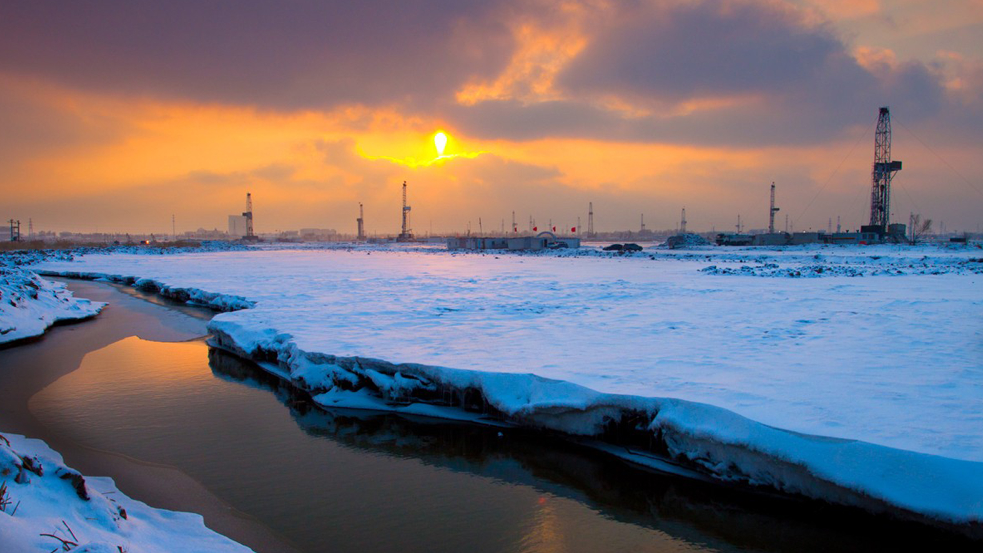 Snow-covered landscape with sunset over oil rigs and a river, creating a serene winter industrial scene.