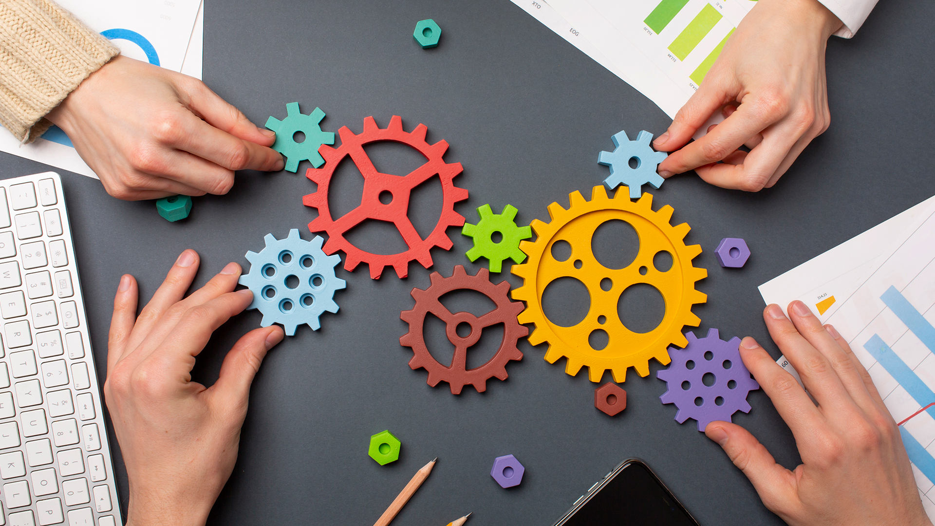 Hands assembling colorful gears on a desk with charts and keyboard, symbolizing teamwork and collaboration.